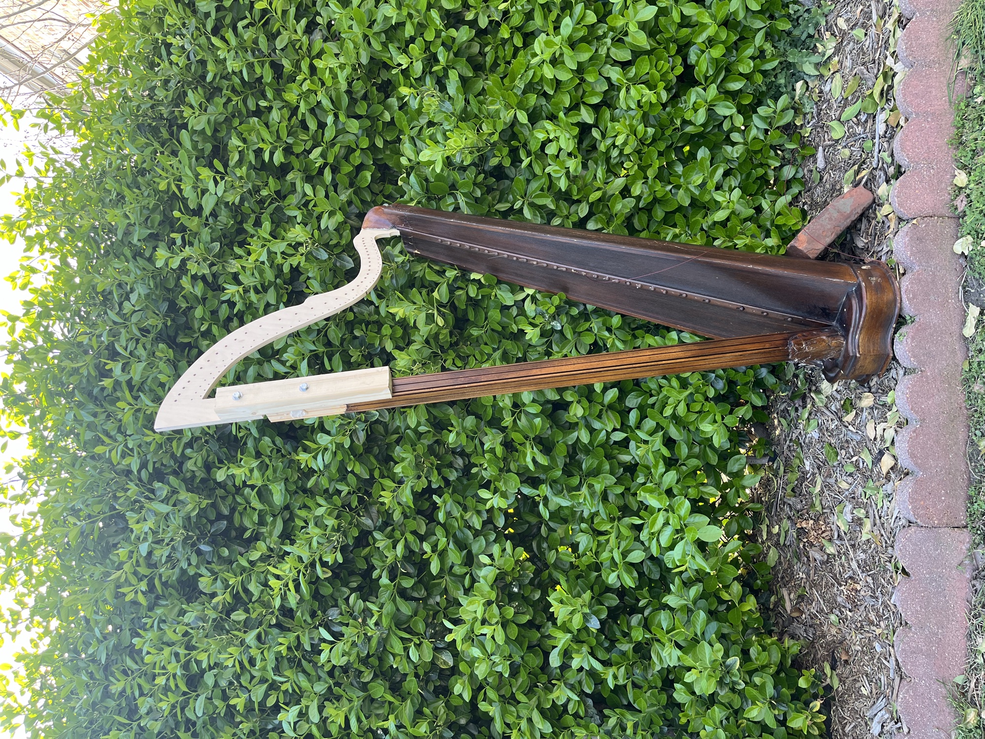 The harp standing outdoors against a hedge, with the pale CNC-cut replacement neck mocked into place on the dark original body.
